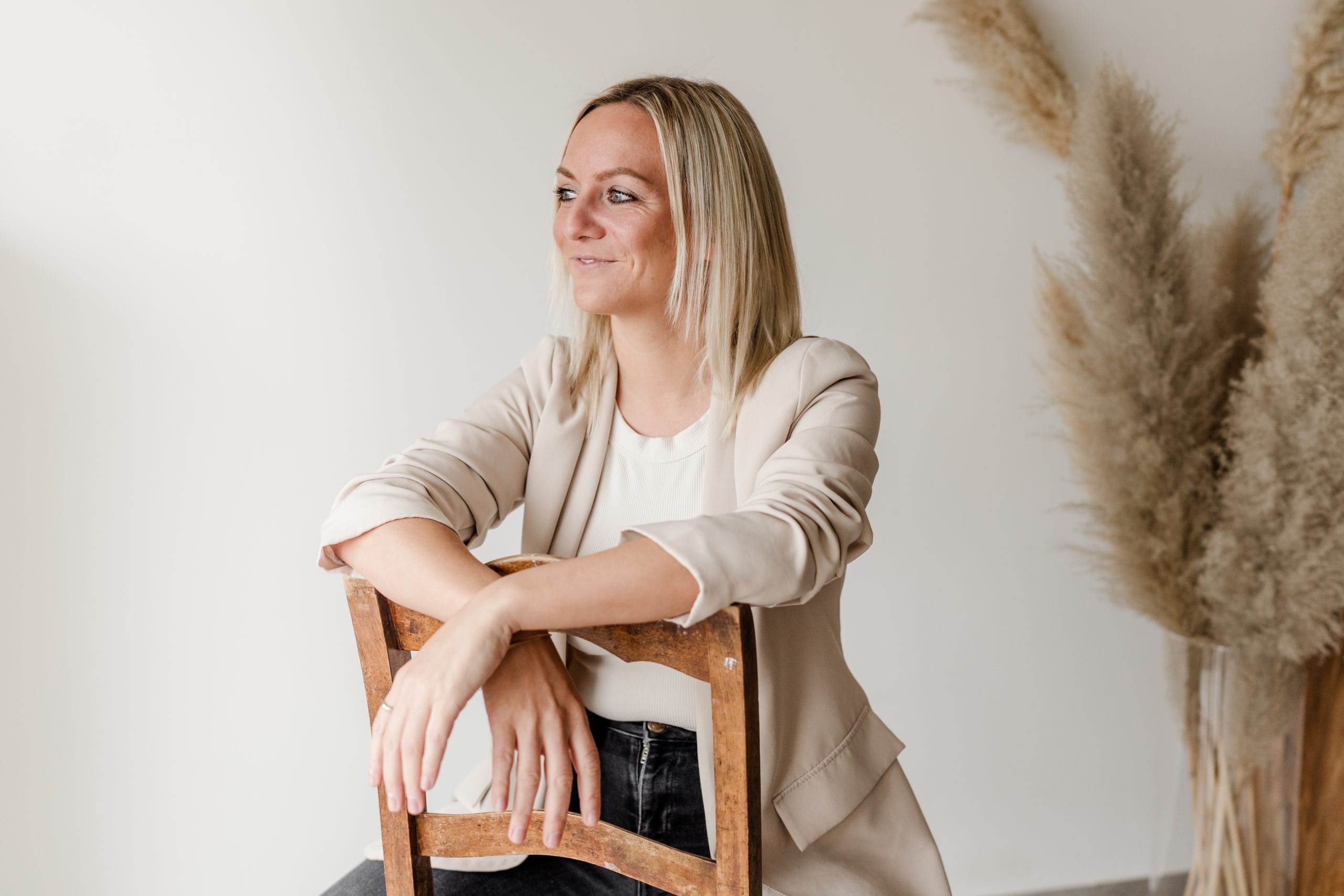 CreativebyJenn Portrait Eine lächelnde Frau sitzt auf einem Stuhl und hat die Arme auf der Lehne überkreuzt.